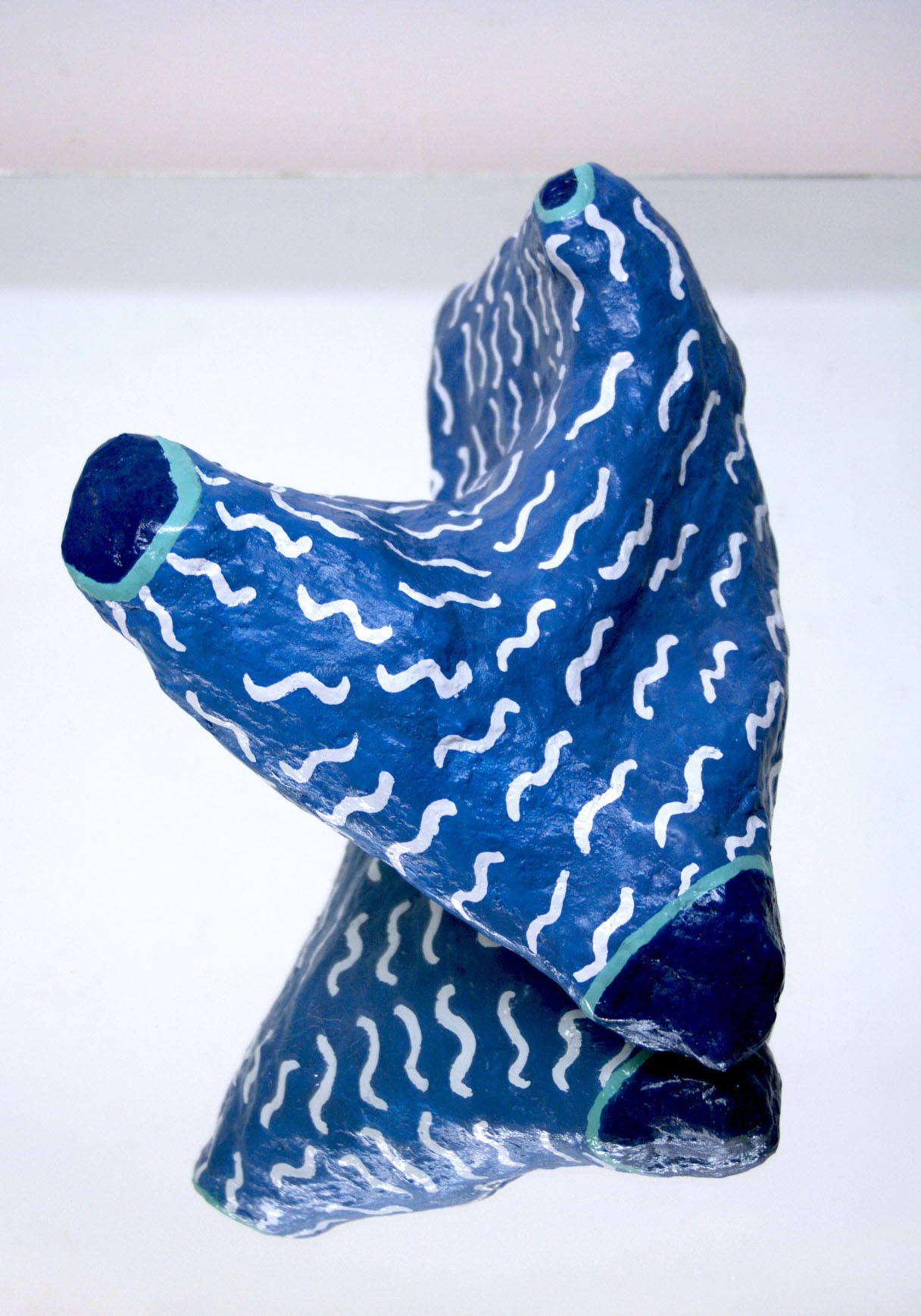 A blue hydrocal abstract shaped sculpture with white squiggly lines and dark blue tips sitting on a mirror